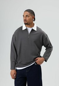 Grijze pullover trui met een witte kraag, halve rits aan de voorkant en lange mouwen, gedragen over een wit overhemd en gecombineerd met donkere spijkerbroek.