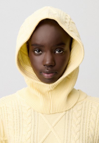 Joven de piel oscura que lleva un suéter con capucha de punto amarillo pálido, mirando directamente al frente con una expresión neutral.