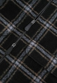 Sort og grå ternet stof med brune detaljer, med lynlås lukning og blødt, tekstureret materiale. Bemærkelsesværdige mønsterdetaljer overalt.