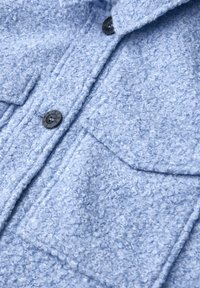 Jasnoniebieska marynarka z teksturowanej tkaniny, wyposażona w dwie przednie kieszenie i zapięcie na guziki. Powierzchnia ma miękki, kręcony wygląd, a guziki są ciemne.