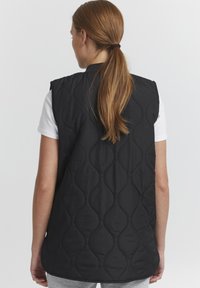 Schwarze wattierte Weste mit ärmellosem Design, ausgestattet mit einem strukturierten, gewellten Muster. Trägt man über einem weißen T-Shirt und kombiniert mit grauen Hosen.