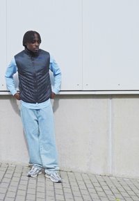 Czarna, pikowana kamizelka na jasnoniebieskiej koszuli z długim rękawem; luźne jasnoniebieskie dżinsy i szare sportowe buty. Stojąc przy jasnej ścianie.