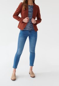 Blazer ajusté couleur rouille porté sur un haut rayé bleu marine et blanc, associé à un jean skinny bleu et des mocassins beige clair. Textures lisses mises en avant.