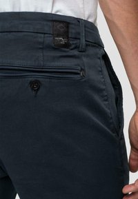 Pantaloni blu navy con tasca con zip e chiusura a bottone, realizzati in tessuto testurizzato con un'etichetta nera brandizzata in vita.