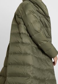Chaqueta parka verde acolchada con cuello alto, textura acolchada y capucha integrada. La tela tiene un acabado suave y un corte holgado.
