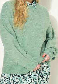 Una persona con lunghi capelli biondi indossa un ampio maglione verde chiaro sopra un vestito a fantasia verde e nera, tenendo il bordo del maglione con unghie smaltate.