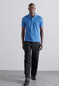 Blå polo skjorte med korte ermer, kombinert med sorte bukser og svarte sko; glatt tekstur og tettsittende design.