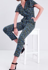 Combinaison en tissu à motifs bleus et verts, avec des manches courtes, un col en V et une taille ceinturée ; ajustée au niveau des jambes, raccourcie au niveau de la cheville.