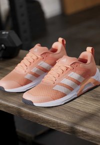 Двойка коралово оранжеви маратонки Adidas с бели ивици и сиви подметки, поставени на дървена повърхност близо до черна дъмбел.
