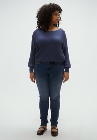 Tengerészkék csíkos hosszú ujjú felső, laza fazonnal, közepes koptatású skinny farmernadrággal és barna balerinacipővel párosítva. A modell oldalt tartja a kezét.