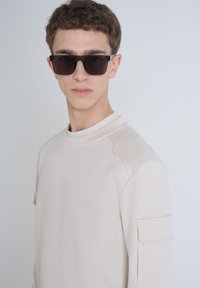 Lichtbeige sweatshirt met een ronde hals, textuurstof en een zijzakdetail op de linker mouw. Donkere zonnebril maakt de look compleet.