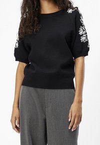 Suéter negro con mangas cortas abullonadas y bordado floral blanco en los hombros. Material de punto suave, cuello y dobladillo acanalados.