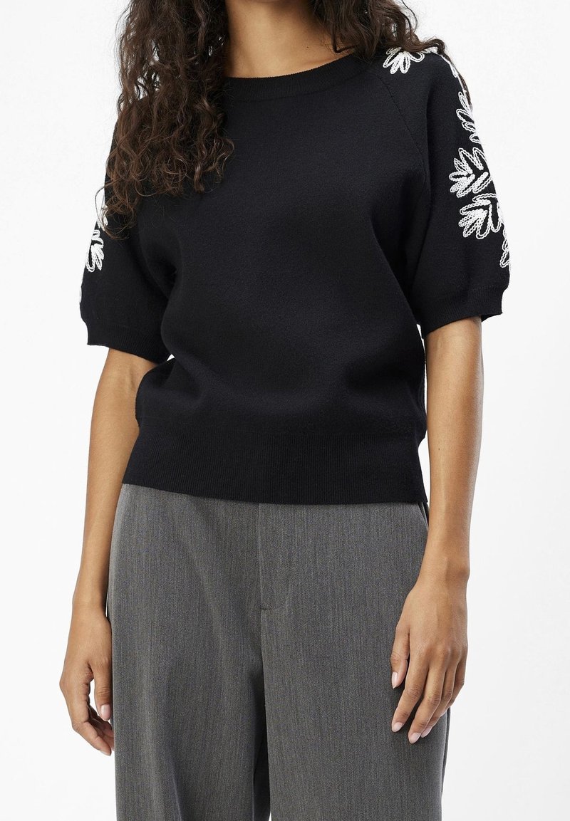 Suéter negro con mangas cortas abullonadas y bordado floral blanco en los hombros. Material de punto suave, cuello y dobladillo acanalados.