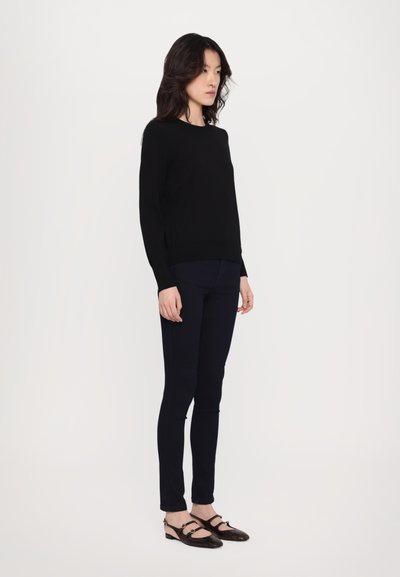 Pulover negru tricotat cu mâneci lungi, asortat cu pantaloni ajustați de culoare albastru închis și pantofi din piele neagră cu curele și accente.