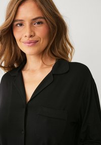 Sort skjorte med knaplukning, krave, korte ærmer og en venstre brystlomme, lavet af et glat, let stof.