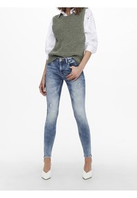 Schlanke blaue Jeans mit leichter Abnutzung, kombiniert mit einem hellgrünen Strickwesten über einem weißen Hemd mit Knöpfen. Weiße High Heels.