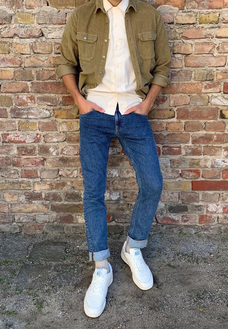 Une personne se tient contre un mur en briques, portant un jean bleu retroussé, des baskets blanches, une chemise beige et une veste vert olive, les mains dans les poches.