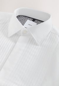 Valkoinen paita, jossa laskostettu etuosa, klassinen kaulus, yksittäinen nappikiinnitys ja kontrastivärinen, kuvioitu kauluksen sisäpinta. Labelissä lukee "NEXT".