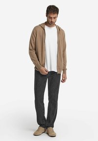 Mann in beigem Zip-up-Hoodie, weißem T-Shirt, dunkelgrauen Jeans und beigen Sneakern, der vor einem schlichten weißen Hintergrund steht.