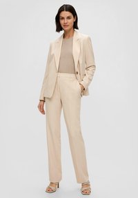 Beige maatpak met een blazer met één knoop en rechte broek. Gedragen over een lichtbeige top. Gladde textuur en minimalistisch ontwerp.