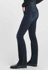 Donker gewassen bootcut jeans met een hoge taille, met achterzakken, subtiele stiksels en een lichte glans. Gecombineerd met glanzende zwarte schoenen.