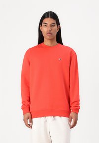 Felpa rossa con maniche lunghe, polsini a coste, scollatura rotonda e piccolo dettaglio del logo sul petto sinistro. Indossata sopra pantaloni chiari.