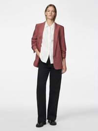 Une femme debout porte un blazer rose poudré, une chemise blanche boutonnée, un pantalon noir à jambes larges et des chaussures noires sur un fond blanc uni.