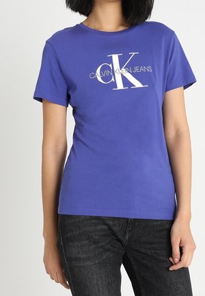 Kvinde iført en lilla Calvin Klein Jeans T-shirt med hvidt logo, kombineret med sorte jeans, stående mod en ensfarvet lys baggrund.