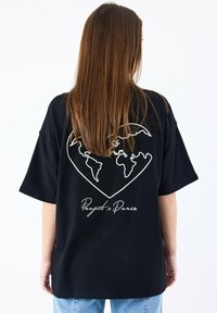 Camiseta negra de gran tamaño con un contorno blanco de un corazón que incluye un mapa del mundo y el texto "Proyecto París" debajo. Manga corta.