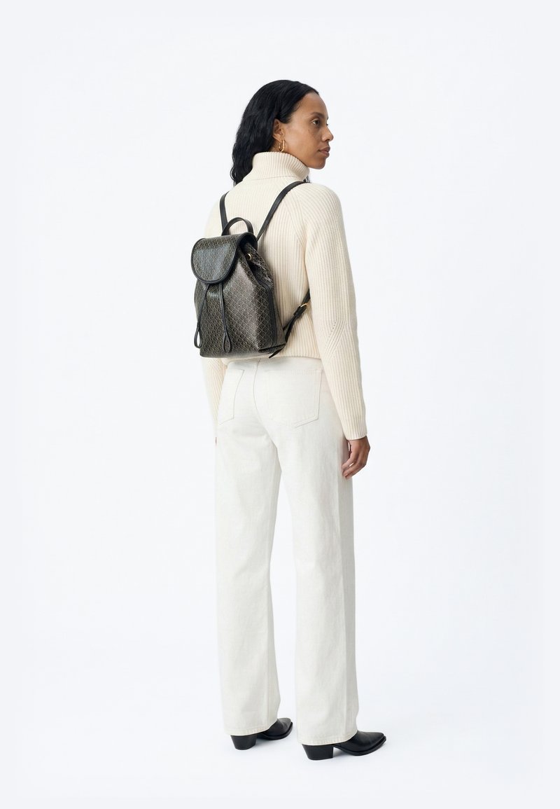 Nő krémszínű garbó pulóvert, fehér bőszárú nadrágot, fekete csizmát visel, és egy sötét mintás bőr hátizsákot hord, oldalirányban áll.