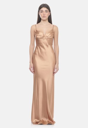 Vrouw in een lange bronzen satijnen jurk met dunne bandjes, een aansluitend bovenstuk en een vloeiende rok, staand voor een effen lichte achtergrond.