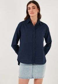 Camicia blu navy con colletto appuntito, maniche lunghe e tessuto morbido e testurizzato. Indossata sopra una gonna di jeans chiaro.