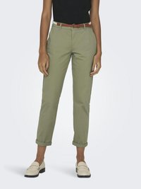 Personne portant un pantalon vert olive retroussé, une ceinture blanche avec une boucle marron, un haut noir et des mocassins crème, debout devant un fond uni.