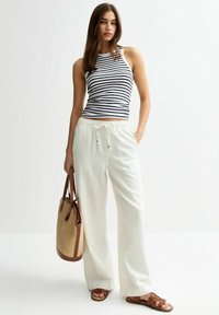 Gestreepte mouwloze top, witte wijde broek, bruine sandalen en een tan geweven totebag. Materiaal lijkt lichtgewicht; ontspannen pasvorm.