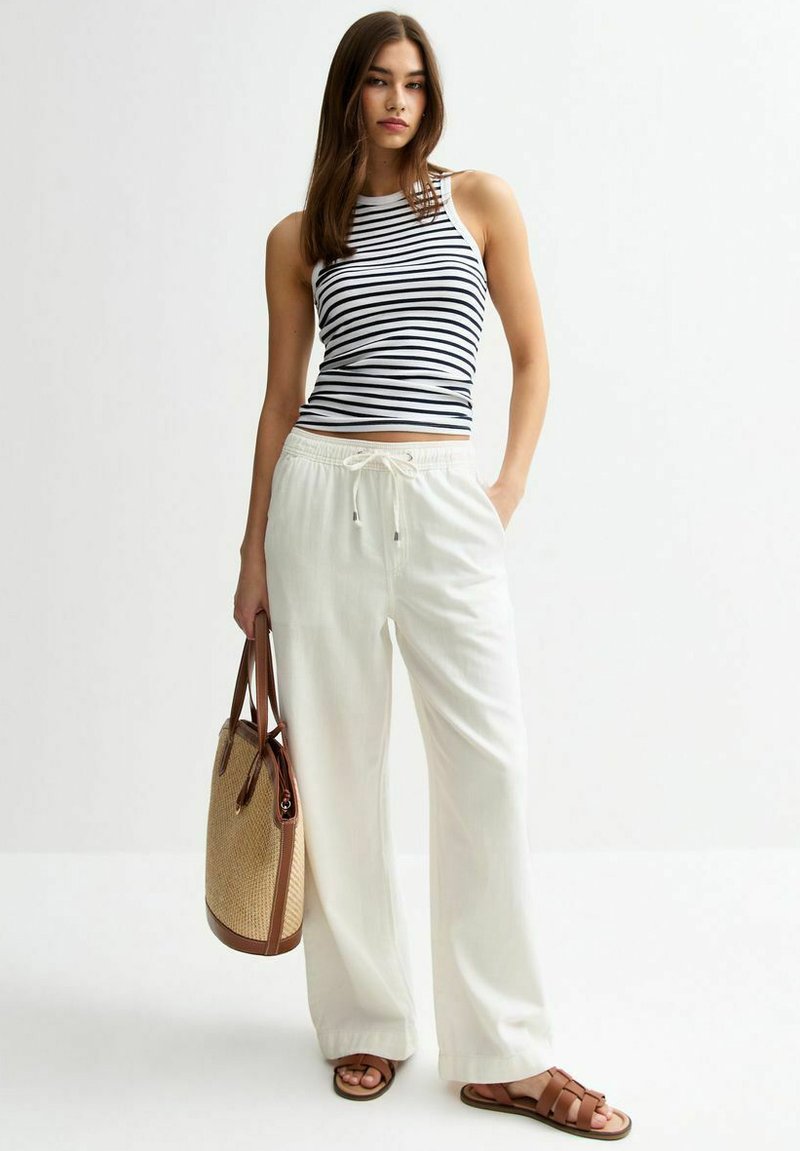 Gestreepte mouwloze top, witte wijde broek, bruine sandalen en een tan geweven totebag. Materiaal lijkt lichtgewicht; ontspannen pasvorm.
