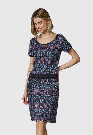 Kvinne iført en kortermet, knelang kjole med fargerike geometriske mønstre og et svart knyttet midjebelte, smilende og ser ned.