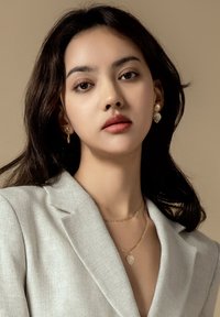Goldene Halskette mit einem Blattanhänger und einer zarten Kette, kombiniert mit passenden Ohrringen in Blattform; das Model trägt einen hellgrauen Blazer.