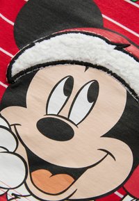 Roter Stoff mit einer großen, gestickten Grafik eines lächelnden Cartoon-Charakters mit schwarzen Ohren und einem flauschigen weißen Hut-Akzent.