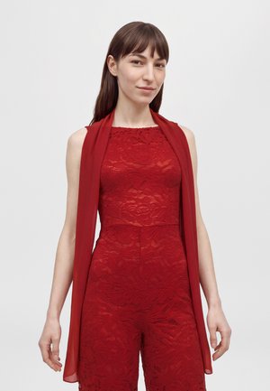 Une femme porte une combinaison-pantalon rouge sans manches en dentelle, accompagnée d’une longue écharpe rouge transparente, sur un fond clair uni.