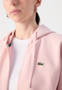 Hoodie zippé rose clair en tissu texturé, arborant un petit logo crocodile vert sur la poitrine gauche et une chemise blanche en dessous.