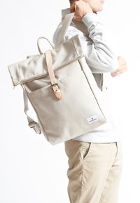 Beige kanvasryggsäck med rulltoppsdesign, läderrem och spänne, framfack och justerbara remmar. Guld dragkedjedetalj.