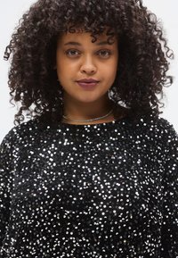 Fekete flitteres felső kerek nyakkivágással, sűrű, tükröződő flitterekkel, amelyek csillogó textúrát alkotnak. Könnyű, lebegős kialakítás.