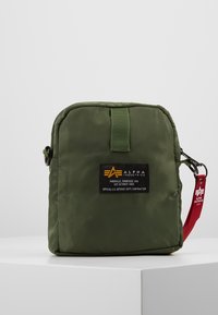 Olivewoblany, nylonowy worek z zamkiem błyskawicznym, z czarną metką z żółtym logo oraz czerwoną akcentującą rączką. Teksturowany, kwadratowy kształt.