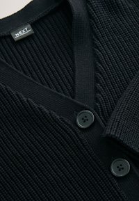 Mornarsko modra pletena jopica z rebrasto teksturo, V-izrezom in dvema črnima gumboma spredaj. Trpežen material s čistim zaključkom.