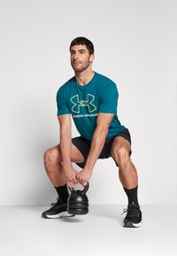 Teal kortærmet skjorte med hvid og gul logo, sorte shorts, holder en sort kettlebell, iført sorte sneakers og sokker.