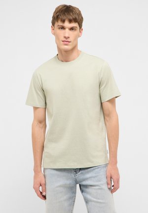 Young man wearing a plain light beige short-sleeve t-shirt and light blue jeans, standing against a plain white background.