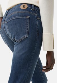 Tmavě modré jeany z denimu s přiléhavým střihem, které mají hnědou koženou nášivku na pase a kontrastní žluté švy na kapsách.