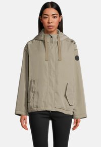 Helle beige Kapuzenjacke aus glattem Material. Mit Reißverschluss, zwei Fronttaschen und einem runden Logo-Patch am Ärmel.