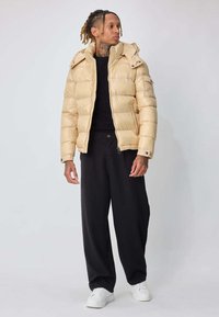 Homme aux dreadlocks portant une doudoune beige, une chemise noire et un pantalon noir, ainsi que des baskets blanches, debout et regardant sur le côté sur un fond blanc.