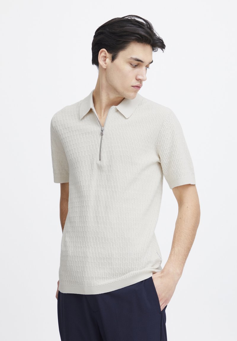 Kortermet beige strikket polo skjorte med et teksturert mønster, glidelås krage og smal passform. Har ribbestrikkede detaljer ved kragen og kanten.